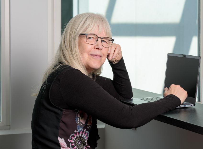 Vi ser Eva-Lena från sidan, hon sitter med datorn och vänder sig mot kameran.